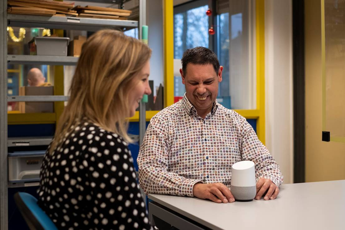 Foto van lachende man, bewoner van Visio wonen met een Google Home speaker. De tafelgenoot lacht ook. Het straalt veel plezier uit.