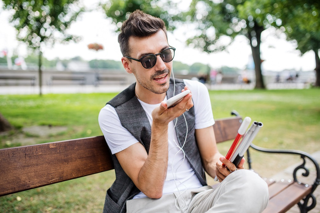Blinde jongen op een bankje in het park. Geeft een spraakopdracht aan z'n telefoon.