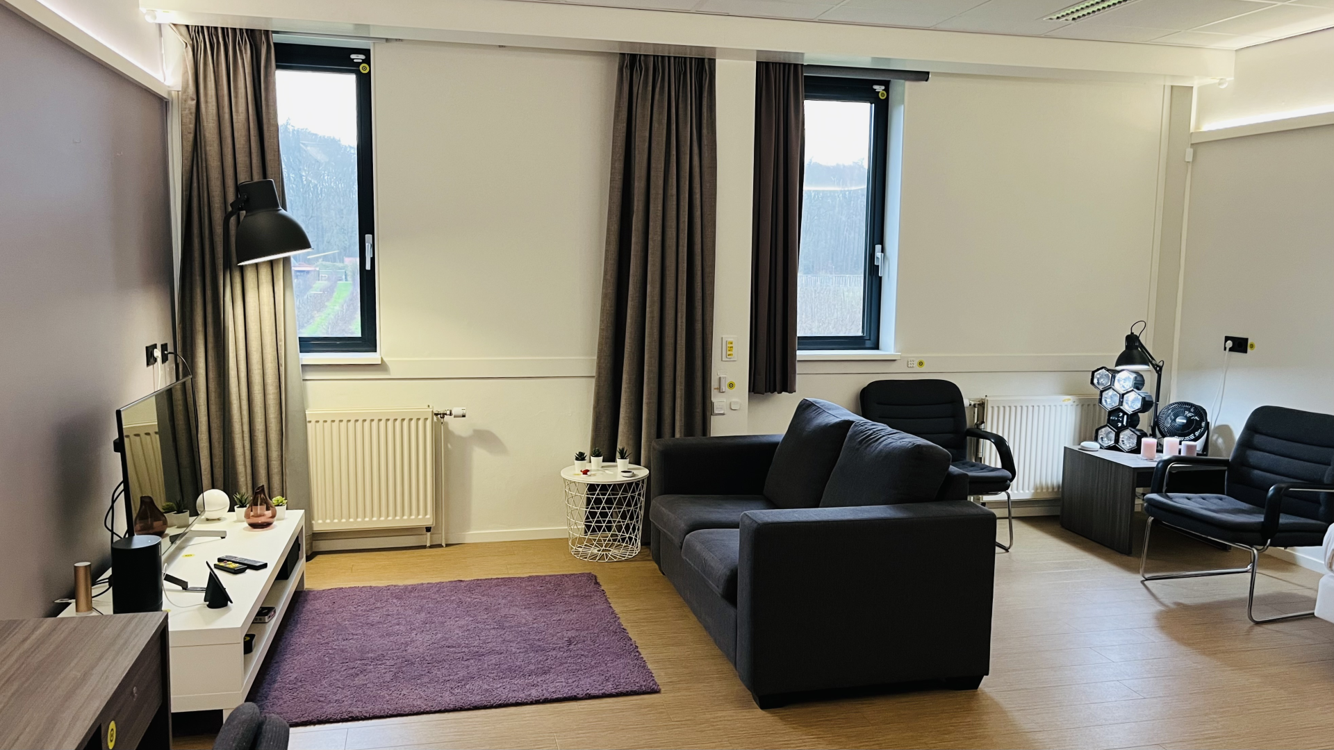 Een overzichtsfoto van De Living Experience Apeldoorn. Je ziet een huiskamer met 2 ramen, gordijnen, een bank, TV, stoel, het valt bijna niet op maar er liggen allemaal slimme snufjes in de kamer.