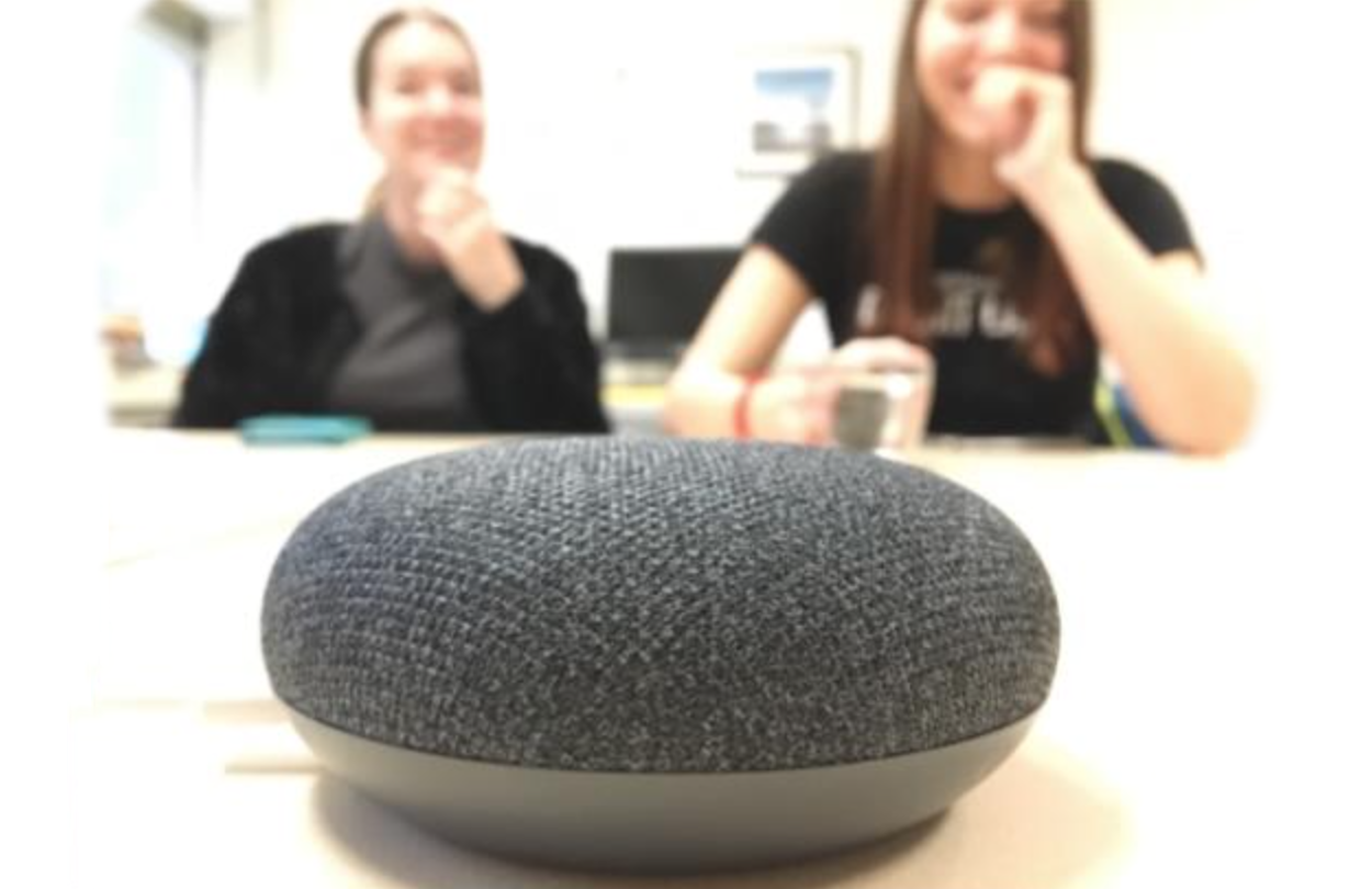 Close-up foto van een Google Nest mini speaker met op de achtergrond 2 lachende revalidanten van Visio Het Loo Erf die meegetest hebben met de VPRO game
