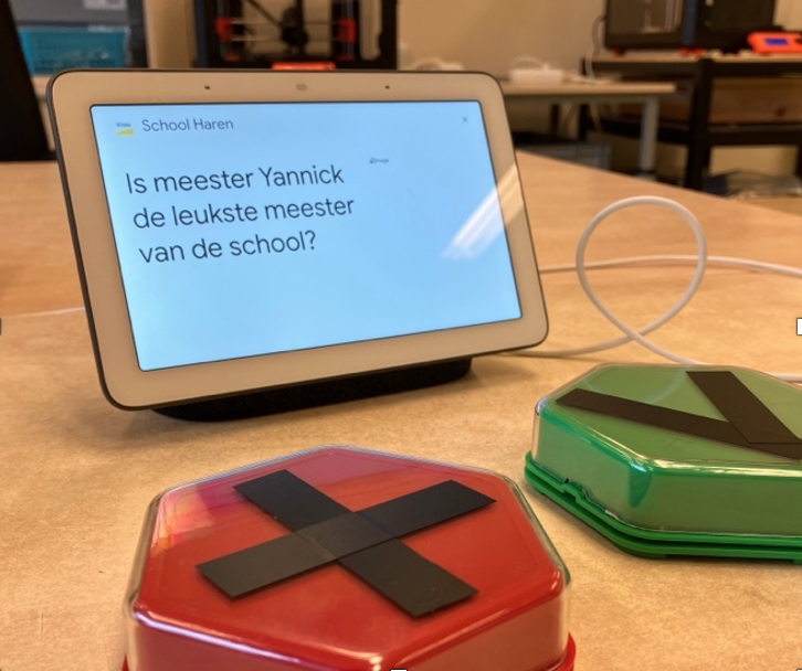 Google nest hub speaker met daarop de tekst "Is meester Yannick de leukste meester van de school?". Voor de speaker liggen 2 knoppen: een rode knop met X en een groene knop met V
